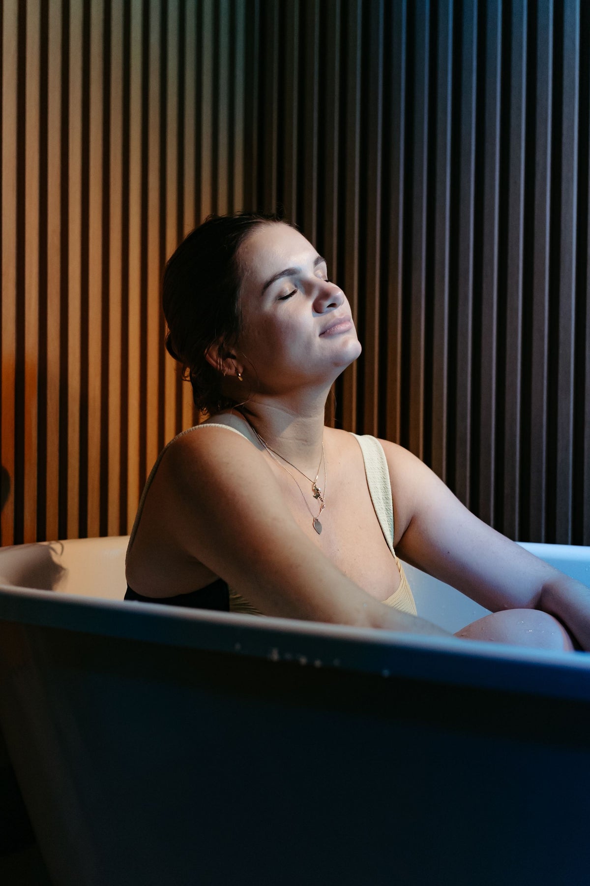 A woman resting with eyes closed in a bath beside dark wood panelling, evoking the quiet of a spa ritual