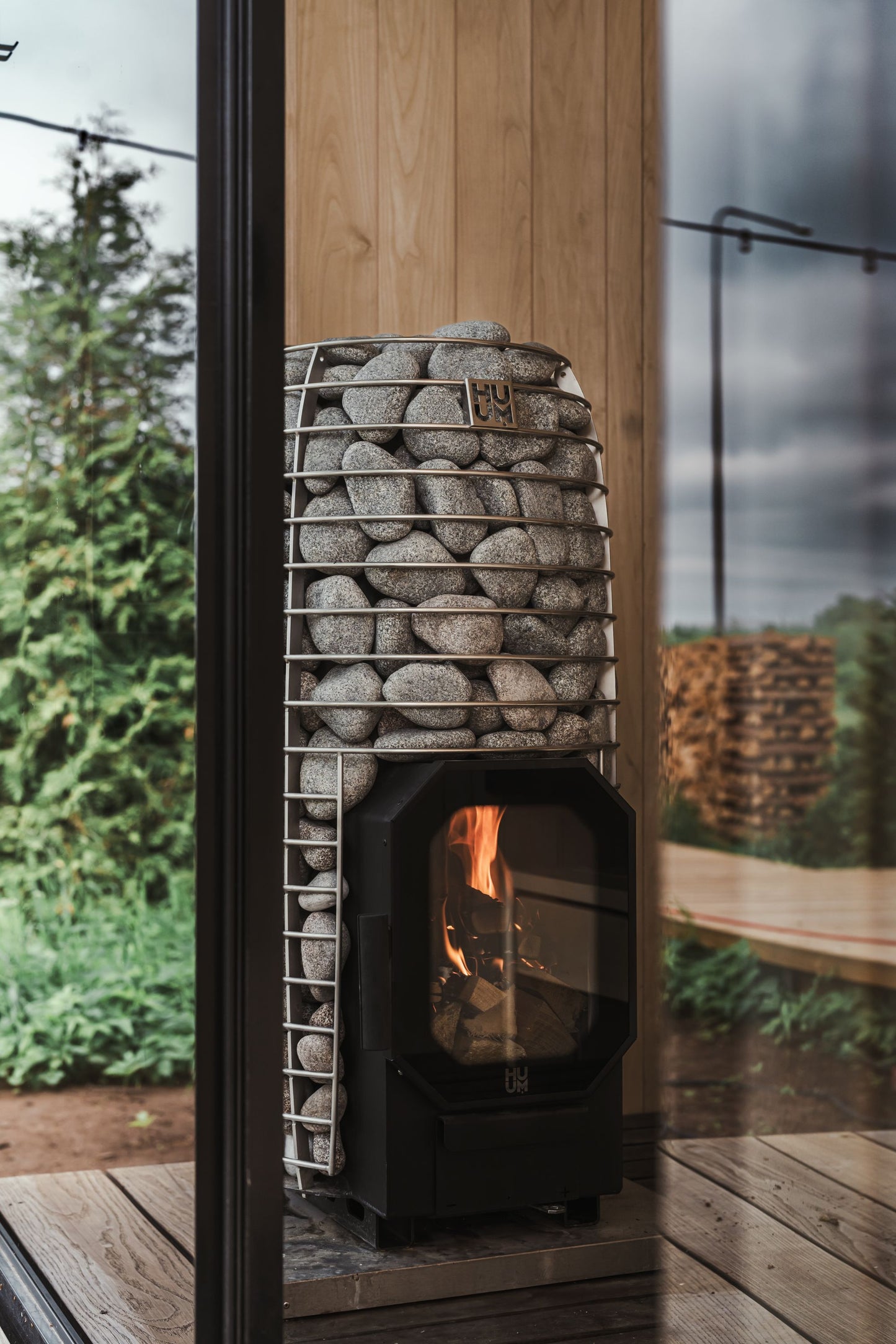 A wood fired stove burning behind stone in a sauna, with trees beyond the glass.