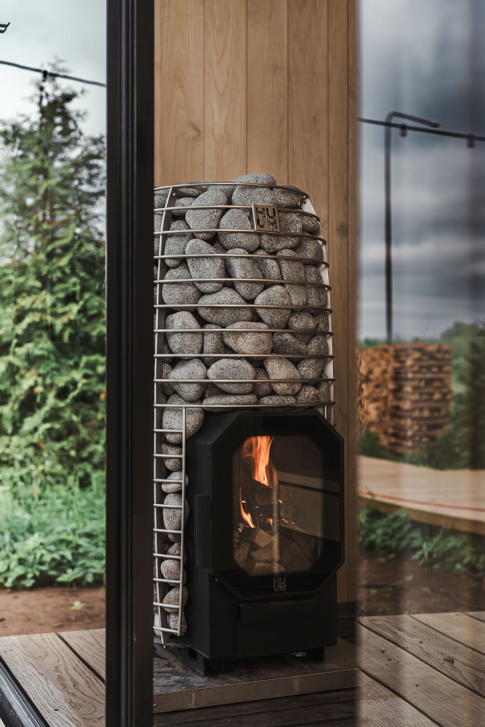 A wood fired stove burning behind stone in a sauna, with trees beyond the glass.