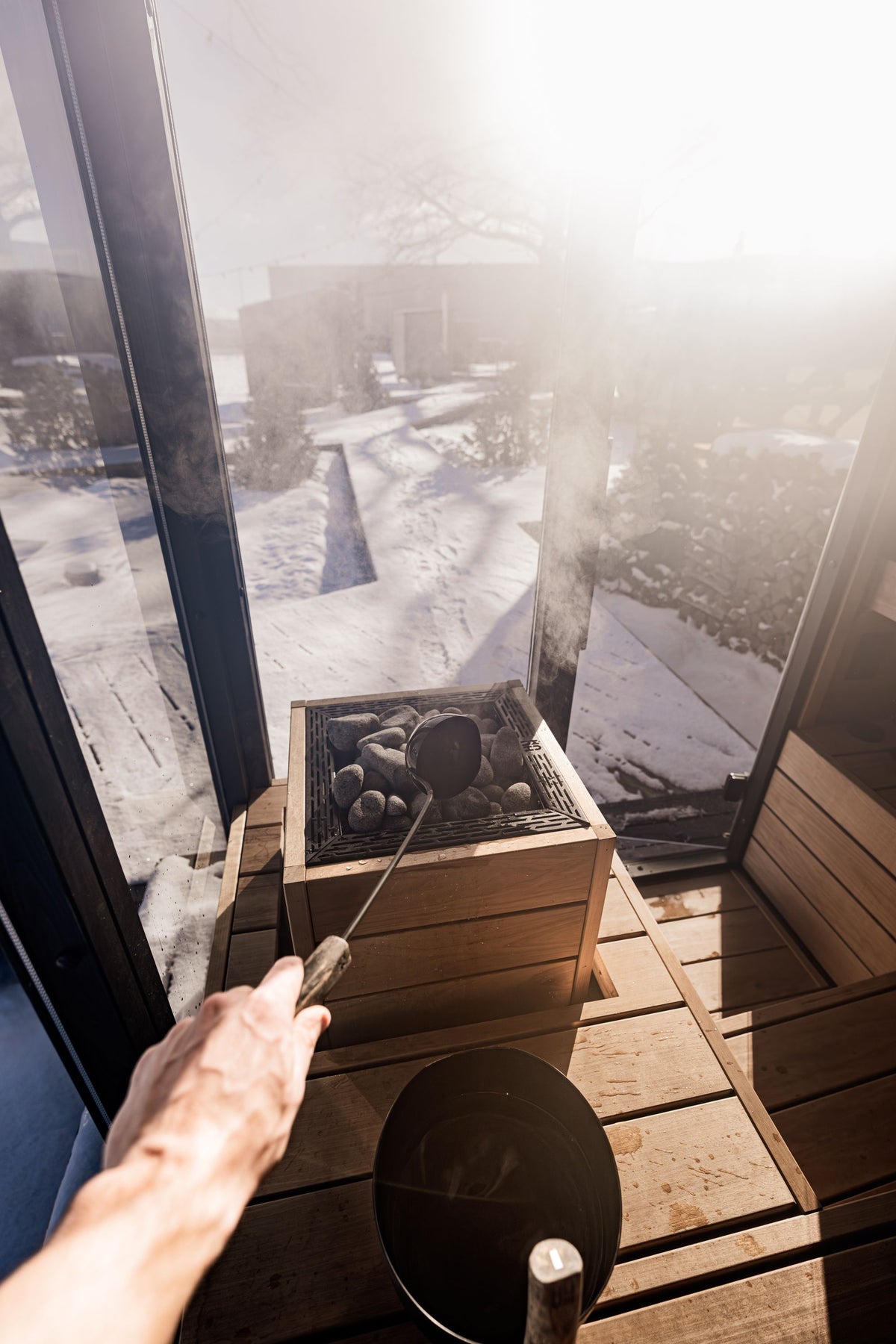 Water ladled onto hot sauna stones as steam glows against the snow beyond the glass, evoking the natural wood sauna at Four Seasons Resort Megève