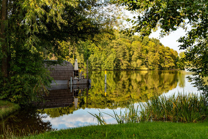 A wooden sauna cabin sits at the edge of a still lake, its reflection perfectly mirrored in the water.