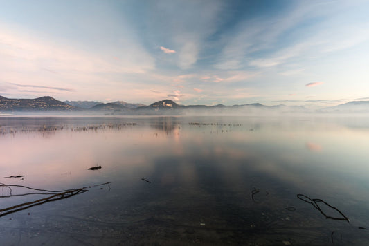 A misty sunrise over a calm lake with mountains in the distance