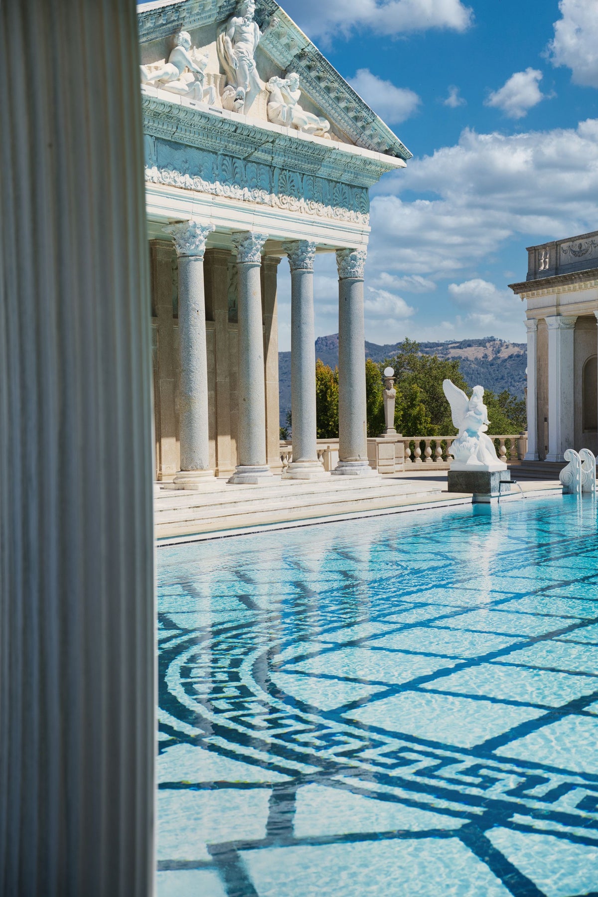 Sunlit marble columns and mosaic tiled water beside a grand classical pool.