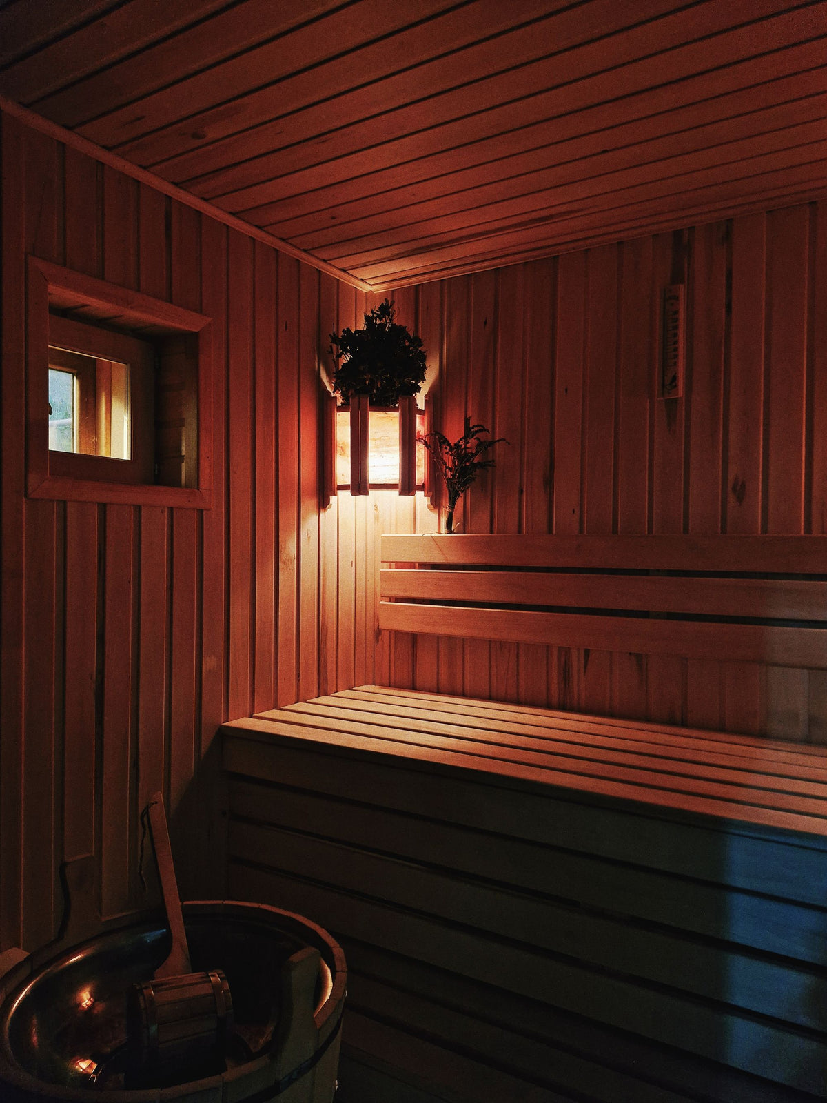 Warm lamplight inside a timber sauna with a small window onto a snowy winter evening