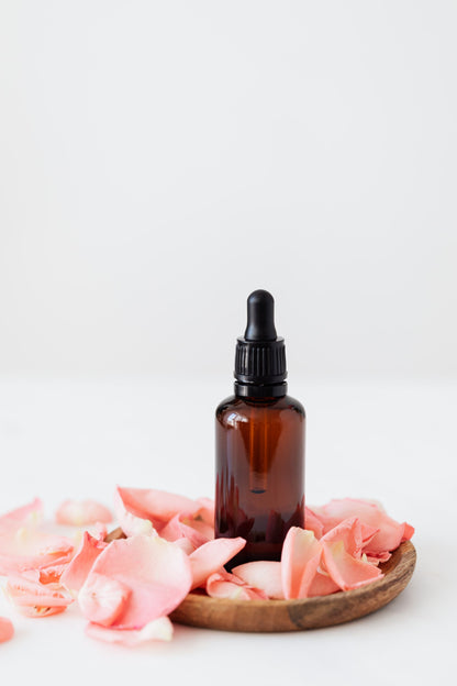 Rose petals and an amber oil bottle set on a wooden plate.