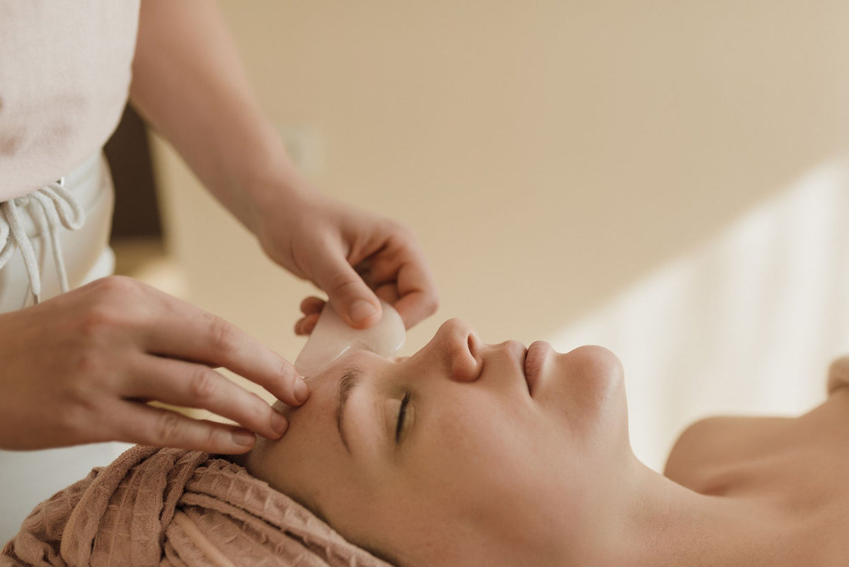 A therapist's hands working a smooth stone across a guest's brow during a facial in warm, soft light, evoking a Dior facial in Paris