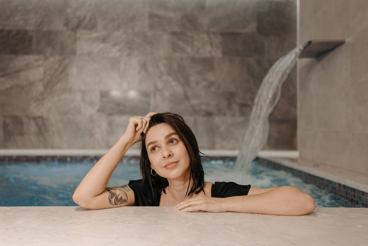 Guest resting at the edge of an indoor pool beside a falling water feature in a stone lined spa