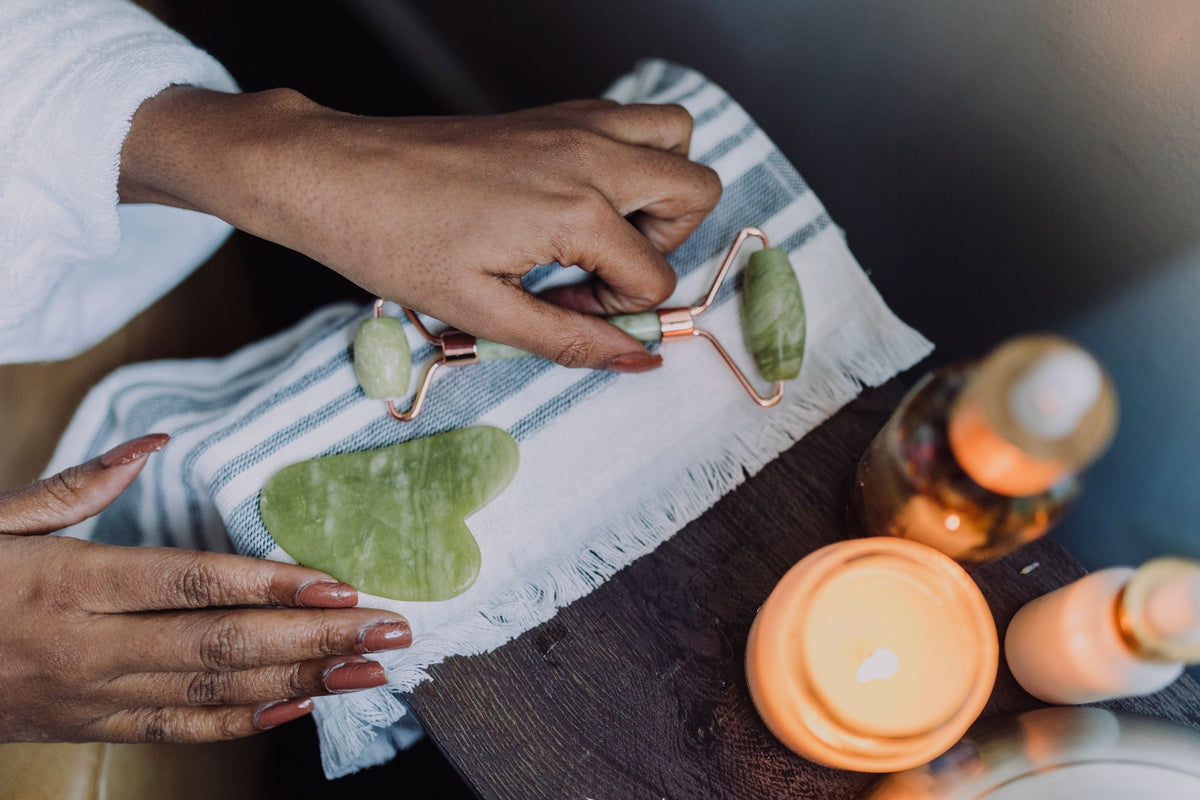 Hands using a jade roller and gua sha stone beside lit candles
