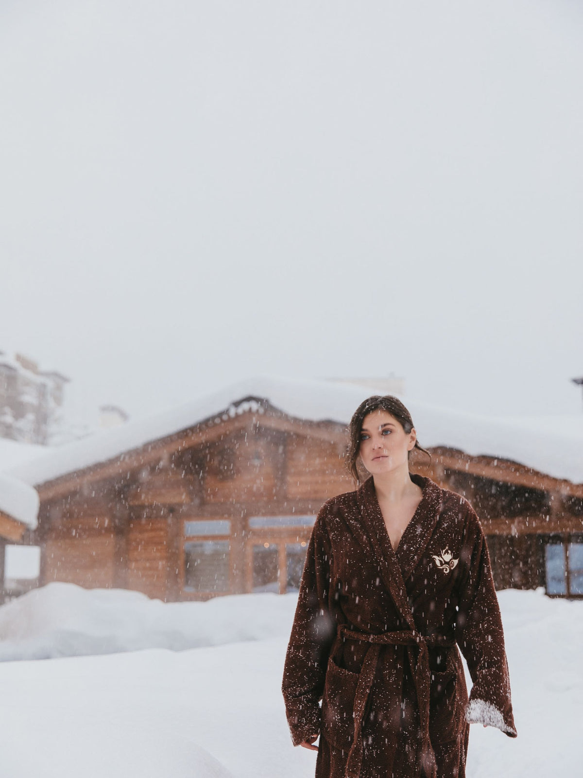 A figure in a spa robe standing in falling snow outside a timber chalet