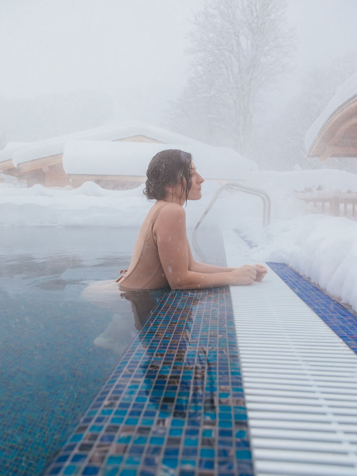 A bather resting in a steaming outdoor pool as snow settles on the chalet roofs behind, evoking the heated pool that runs from indoors to out at Four Seasons Resort Megève