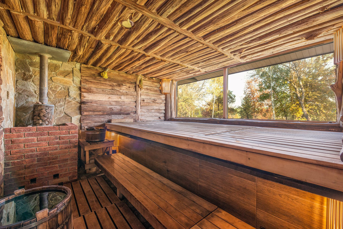 A rustic wood lined sauna with a stone stove looks out through a window to the trees beyond.