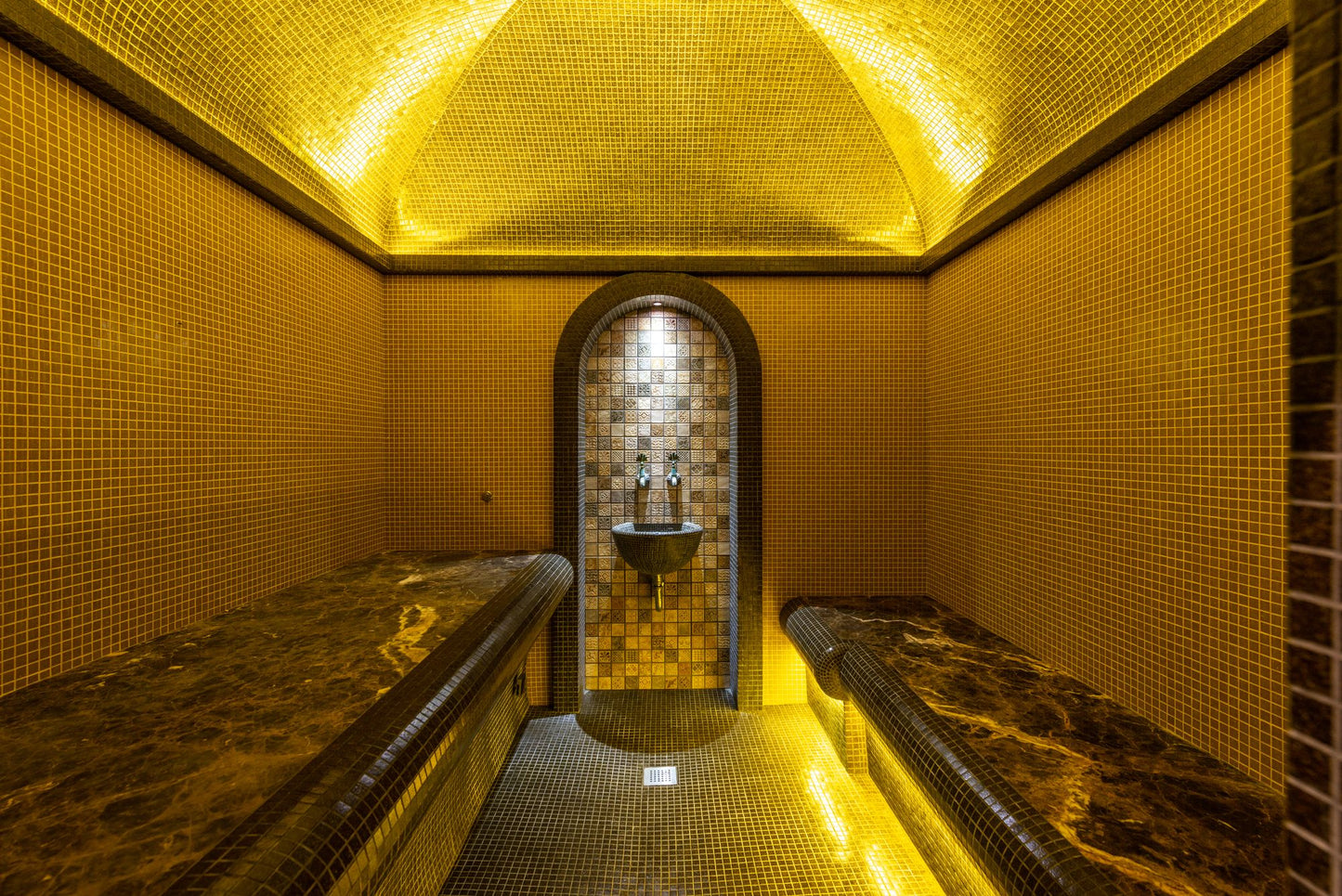 A small steam chamber lined floor to vault in gold mosaic with dark marble benches, standing for the marble hammam suite
