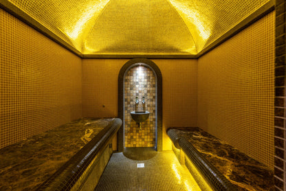 A small steam chamber lined floor to vault in gold mosaic with dark marble benches, standing for the marble hammam suite