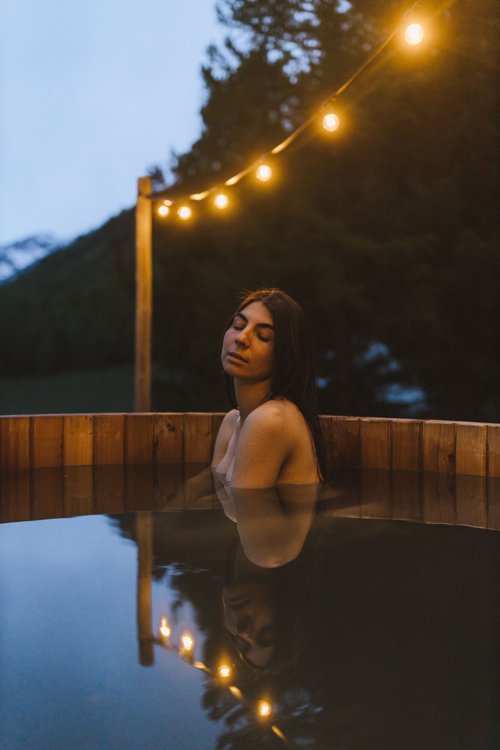A woman bathing in a wooden tub at dusk under a string of warm lights, The Cowshed Spa at Soho Farmhouse