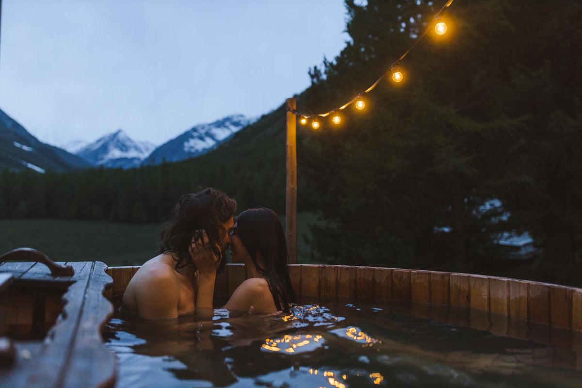 A couple relaxing together in an outdoor hot tub at dusk with snow capped Alps behind them