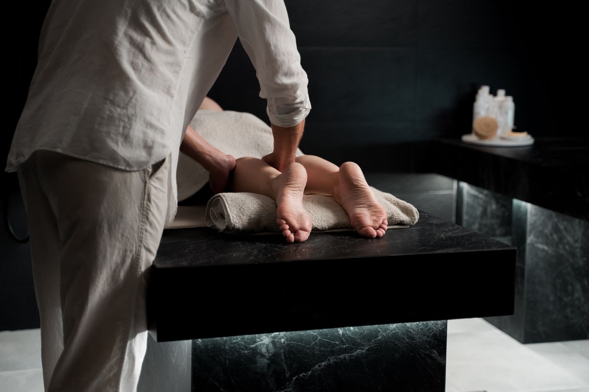 A therapist working with both hands on a lower leg against dark marble, the register of the treatment rooms
